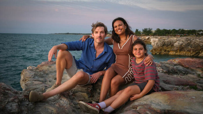 Gwilym Lee, Miranda Tapsell and Gladys-May Kelly sit on a rock by the ocean.